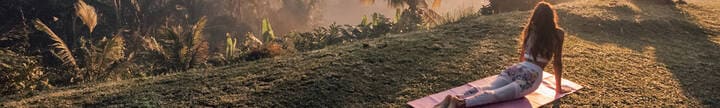 A woman performs yoga poses on a mat set on lush green grass, overlooking serene rice fields as the sun rises.