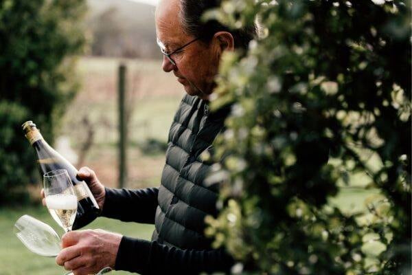 A man standing behind a tree in Tasmania's Tamar Valley holds a bottle of sparkling wine and champagne flutes.