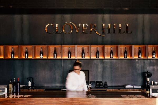 A staff member at Clover Hill Restaurant is seen working behind the bar, skillfully preparing drink orders for customers. The bar area features an array of bottles and glassware, creating a welcoming atmosphere as the staff member focuses on crafting beverages.