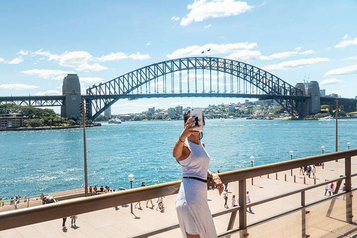 Woman taking selfie with mobile phone at iconic Sydney Harbor Bridge. Cityscape, water, with buildings in CBD area. Tourist attraction in Australia. Technology, tourism, travel, taking a photo concept
