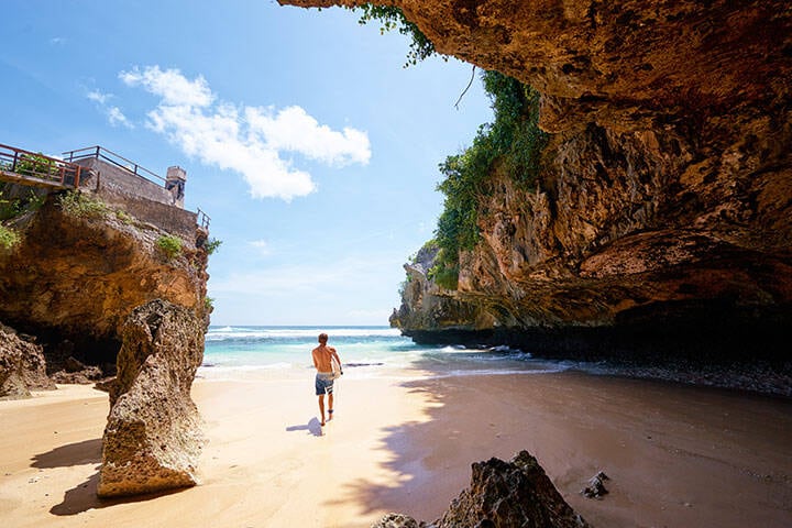 Surfer holding board walking on sand at bottom of beach in Uluwatu, Bali